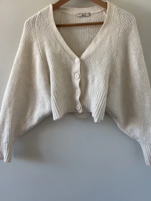 Vit ribbad kofta från Gina tricot  - Säljer en krämvit ribbstickad kofta från Gina tricot. Koftan har v-ringning, stora vita knappar framtill och en croppad passform. 