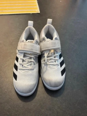 Vita Adidas sneakers, powerlift 5 -  Säljer ett par vita adidas Powerlift 5 i storlek 40. Perfekta för gymmet – ger bra stabilitet och stöd vid t.ex. knäböj och annan styrketräning. Fast sula och bra grepp som hjälper dig stå stadigt under lyft. Snygga vita Adidas sneakers med klassiska svarta ränder på sidan. Skorna har både snörning och kardborreband. Har använt dom men dom är fortfarande i fin funktion. Den enda anmärkningen är att dom blivit lite smutsiga  Perfekta om du vill testa lyftarskor utan att köpa nya. Nypris ca 1200 