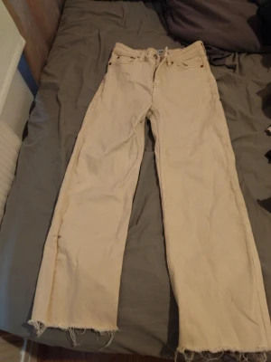 Beige raka jeans med rå kant - Säljer ett par beige jeans med rak passform och råa, fransiga benslut. Jeansen har klassisk femficksdesign och knappgylf. Perfekta för en avslappnad och trendig look.
