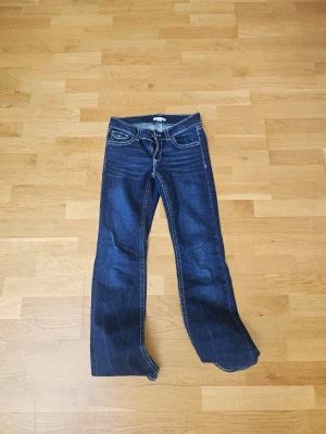 Gina young mörkblåa jeans med fickor - Snygga mörkblå bootcut jeans med vita kontrastsömmar och klassiska fem fickor. Jeansen har dekorativa ficklock bak och lätt slitna detaljer framtill. Perfekta för en avslappnad och trendig look.