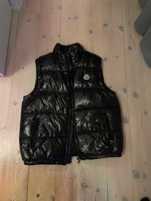 Svart dunväst från Moncler - Snygg svart dunväst från Moncler med glansig finish och hög krage. Västen har två dragkedjeförsedda fickor och klassisk Moncler-logga på bröstet. Perfekt för lager-på-lager och ger en clean streetwear-look.