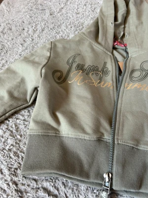 Olivgrön hoodie med tryck och dragkedja - Snygg olivgrön hoodie från Jump Style med huva och dragkedja framtill. Hoodien har ett coolt tryck i svart och guld på bröstet och breda ribbade muddar vid midjan. Perfekt för en avslappnad och trendig look.