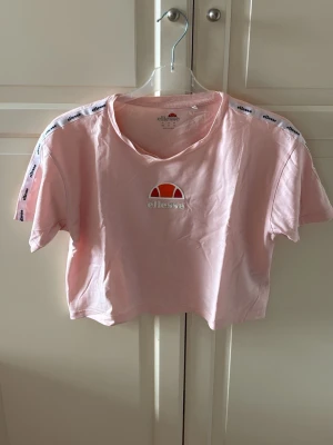 Rosa croppad t-shirt från Ellesse - Snygg ljusrosa croppad t-shirt från Ellesse med logga framtill och logoband längs axlarna. Klassisk rund hals och korta ärmar. Storlek 40. I fint skick. 