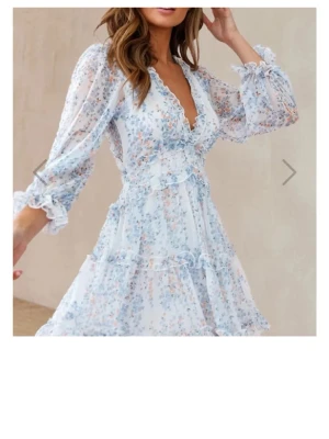 Somrig, blommig långklänning med volanger, XS🌸  - Romantisk långklänning i vitt med blått och ljusrosa blommönster. Klänningen har v-ring, volangdetaljer, transparenta långärmar och öppen rygg. Tillverkad i lätt och luftig polyester, perfekt för sommarens alla tillfällen🌸! 