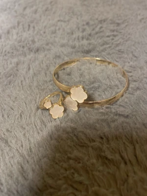 Guldigt armband med vita blommor - Snyggt armband i guldfärgad metall med två vita blommor i pärlemor-look. Blommorna har en mjuk, rundad form och sitter i ändarna på armbandet. Perfekt accessoar för dig som gillar en söt och trendig stil.