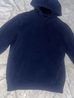 Mörkblå hoodie med huva - En enkel och stilren mörkblå hoodie med huva och dragsnören. Hoodien har en klassisk passform, känguruficka framtill och ribbade muddar vid ärmslut och nederkant. Perfekt för en avslappnad look och lätt att matcha med jeans eller joggers.