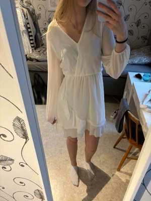 Vit omlottklänning med volang - Söt vit kortklänning med omlott och v-ringning. Klänningen har långa, transparenta ärmar och volangdetaljer nertill. Har även ett knytband i ryggen🫶🏻