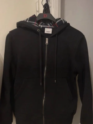 Svart hoodie med dragkedja och luva - Klassisk svart hoodie från Burberry med hel dragkedja framtill, justerbar luva med snören och känguruficka. Insidan av luvan har det ikoniska Burberry-rutiga mönstret. Perfekt för en avslappnad och stilren look. 