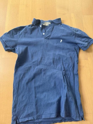 Marinblå pikétröja med krage - Snygg marinblå pikétröja med klassisk krage och två knappar framtill. Tröjan har korta ärmar och en liten vit logga på bröstet. Perfekt till jeans eller shorts för en avslappnad look.