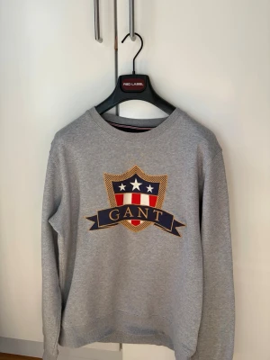 GANT: Shield Sweatshirt  - Snygg grå sweatshirt från GANT med stort tryck framtill i form av ett sköldmärke med stjärnor och ränder i blått, vitt och rött. Tröjan har rund halsringning och ribbade muddar vid ärmslut och nederkant. Perfekt för en avslappnad och stilren look både med ett par chinos men även jeans! 🎉För mer info skriv dm!