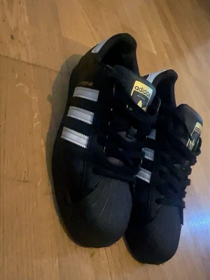 Adidas Superstar svarta sneakers - Den här modellen är en riktig klassiker och otroligt populär världen över – den har burits av generationer och är fortfarande en streetwear-favorit. Kändisar som Marlon (och många andra) har haft exakt samma stil på sig, vilket gör dem extra eftertraktade just nu. Skorna ser grymma ut både till vardagsjeans, chinos eller ett street-look. Kommer i originalskick utan några defekter eller slitage. Vill du ha ett par riktiga ikoner som känns lyxiga men samtidigt slitstarka? Då är det här dina!