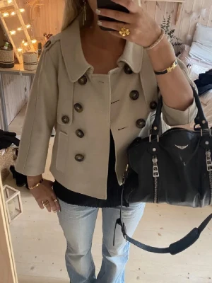 Kort beige dubbelknäppt jacka - Snygg kort jacka i beige med dubbelknäppning och stora bruna knappar. Jackan har trekvartsärmar, bred krage och två fickor framtill. Materialet är 100% ull