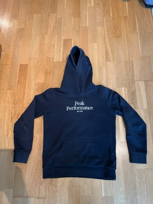 Peak Performance hoodie - Säljer nu denna snygga svarta hoodie från Peak Performance. Den är i storlek 160 och har en ganska slimmad passform. Den har tecken på användning och är lite urtvättad. Utöver det är den i fint skick. Vid fler bilder eller frågor är det bara att höra av sig!