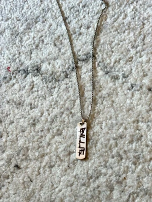 Billie Eilish Billie Silverfärgat halsband med bricka - Silvrigt Billie Eilish halsband. Kedja är cirka 60cm. Ny. Rostfritt stål. Snyggt halsband i silverfärg med en rektangulär bricka som har svart text på framsidan. Kedjan är tunn och har ett klassiskt lås. Perfekt accessoar för dig som gillar stilrena och personliga smycken.