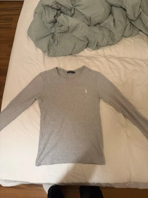 Grå långärmad tröja från Ralph Lauren - Snygg grå långärmad tröja från Ralph Lauren med rund halsringning och diskret broderad logga på bröstet. Tröjan är i mjukt material och har en klassisk, slim fit passform som funkar till många olika stilar.