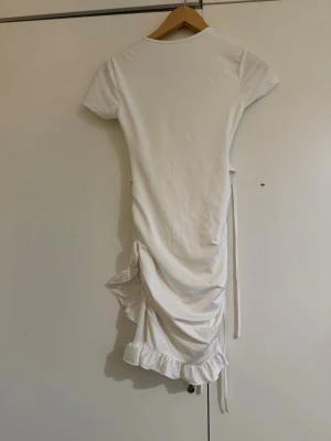 Vit kort klänning med snörning - Supersöt vit kortklänning med volangkant och snörning framtill. Klänningen har korta ärmar, omlott-detaljer och rynkade sidor som ger en snygg passform. Perfekt för sommarens alla tillfällen!