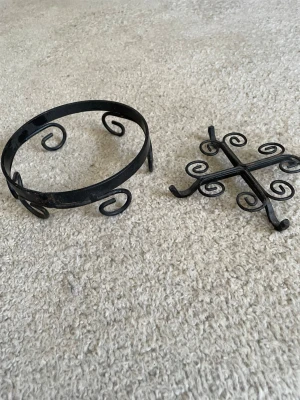 Svart smidesunderlägg i metall - Runt underlägg i svart smide med dekorativa snirkliga detaljer. Perfekt att ställa kastruller eller grytor på för att skydda bordet. Tillverkat i metall och har en klassisk, rustik känsla. I två delar kan användas var för sej 