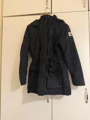 Svart parkasjacka med huva - Säljer en svart parkasjacka med huva och dragsko i midjan. Jackan har flera fickor framtill, patch på ärmen och dragkedja med vindslå. Perfekt för kyliga dagar och har en stilren, längre passform och sitter väldigt fint på.