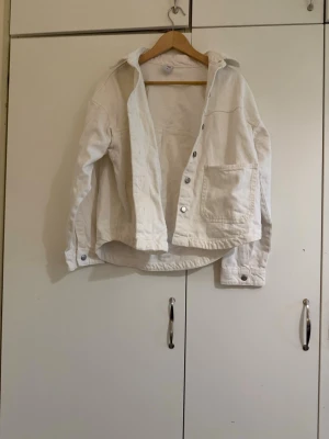 Vit jeansjacka - Snygg vit overshirt i avslappnad passform med stora silverfärgade knappar och en bröstficka. Overshirten har klassisk krage och långa ärmar med knapp vid ärmslut. Perfekt att slänga över en t-shirt för en cool look.