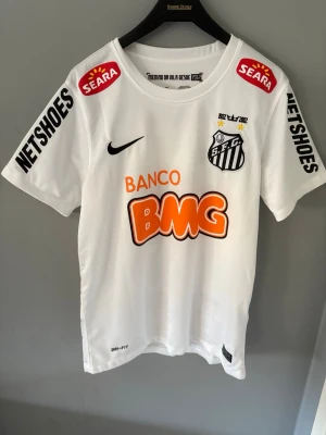 Vit Santos FC Neymar Jr matchtröja - Säljer en vit Santos FC matchtröja från Nike med Neymar Jr och nummer 11 på ryggen. Tröjan har flera sponsorloggor i svart, orange och rött samt klubbmärke på bröstet. Tillverkad i lätt och ventilerande Dri-FIT material, perfekt för fotbollsträning eller match.