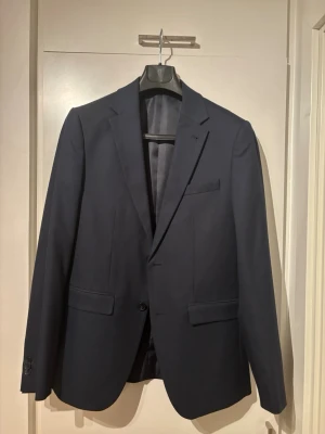 Mörkblå klassisk kavaj herr - Stilren mörkblå kavaj med två knappar framtill, två fickor med lock och en bröstficka. Kavajen har smal passform och klassiska slag. Perfekt för en dressad look. Materialet ser ut att vara en slät vävd textil, troligen ullblandning. Slim fit. Använd en gång.