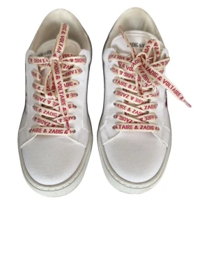 Zadig & Voltaire vita sneakers - Vita sneakers från Zadig & Voltaire med röda broderade läppar på sidan och textdetaljer. Skorna har vita sulor och vita snören med röd logotyptext. Tillverkade i tyg och läder för en cool och trendig look. Nypris ~3000-4000kr. Gå in på min vinted för att köpa ett pris för 2100kr😄