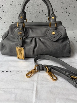 Grå handväska från Marc by Marc Jacobs - Snygg grå handväska i mjukt läder från Marc by Marc Jacobs. Väskan har guldfärgade metalldetaljer, dubbla handtag och avtagbar axelrem. Dekorativ metallbricka framtill och fina sömmar ger en exklusiv känsla. Perfekt för dig som gillar stilrena accessoarer.