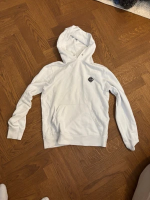 Vit hoodie med patch från J Lindeberg - Säljer en stilren vit hoodie från Svea med en svartvit patch på bröstet. Hoodien har klassisk huva med dragsko och en stor magficka framtill. Tillverkad i mjukt bomullsmaterial som är skönt att bära. Perfekt för dig som gillar enkel och clean streetwear.