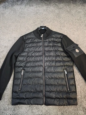 Svart dunväst från Moncler - Cool svart dunväst från Moncler med quiltad framsida och stickade ärmar. Västen har två dragkedjefickor fram och en snygg Moncler-logga på ärmen. Materialet är en mix av dun och stickad textil, perfekt för att hålla värmen. Stängs med dragkedja och har ribbad krage.