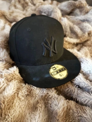 Svart New Era keps - Säljer en svart New Era 59FIFTY keps med NY Yankees-logga i svart brodyr på framsidan. Klassisk flat brim och strukturerad form. Kepsen är gjord i bomull och har en guld etikett på skärmen. Perfekt för dig som gillar streetstyle.