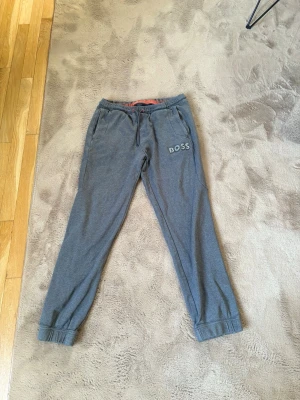 Grå mjukisbyxor från BOSS - Snygga grå mjukisbyxor från BOSS med resår i midjan och benslut. Byxorna har två sidofickor och en dragsko i midjan för skön passform. BOSS-logga broderad på vänster ben. Perfekta för chill eller träning.