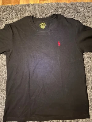 Svart t-shirt från Polo Ralph Lauren - Klassisk svart t-shirt från Polo Ralph Lauren med röd broderad logga på bröstet. T-shirten har rund halsringning och korta ärmar. Tillverkad i mjuk bomull för skön känsla och enkel stil.