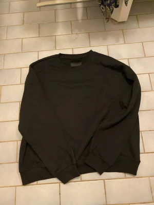 Svart sweatshirt från 157 - En enkel och stilren svart sweatshirt från 157 med rund halsringning och ribbade muddar vid ärmslut och nederkant. Perfekt basplagg för alla tillfällen och lätt att matcha med andra kläder.