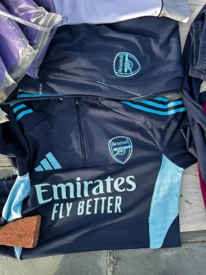 Mörkblå Arsenal träningsjacka Adidas - Snygg mörkblå träningsjacka från Adidas x Arsenal med ljusblå detaljer och klubbmärke på bröstet. Jackan har halv dragkedja, tryckta loggor och texten 'Emirates Fly Better' framtill. Perfekt för fotbollsträning eller sportig stil.