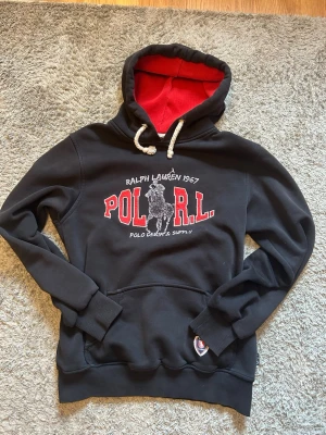  Ralph Lauren vintage hoodie - Säljer en svart hoodie från Polo Ralph Lauren med röd insida i huvan och vita dragsnören. Stor broderad logga och text på bröstet i rött och vitt. Klassisk känguruficka framtill och ribbade muddar. Perfekt för en avslappnad och sportig stil.