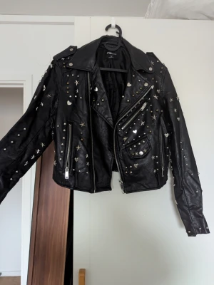 Svart skinnjacka med nitar och stjärnor - Svart skinnjacka i fauxläder med silvriga nitar, hjärtan och stjärnor över hela jackan. Jackan saknar några nitar och på kragen vid insidan av jackan finns slitage (se bilder). 