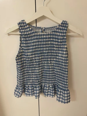 Blåvit rutig ärmlös topp - Söt ärmlös topp med blåvitt rutigt mönster och smockad struktur. Toppen har volangkant nedtill och rund halsringning. 
