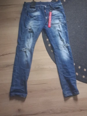 Blå slitna jeans från Dsquared2 - Säljer ett par blå jeans från Dsquared2 med slitningar och distressed detaljer. Jeansen har en smal passform och klassisk femficksdesign. Materialet är denim och det finns en röd Dsquared2-tag vid gylfen samt en stor röd nyckelring med logga.