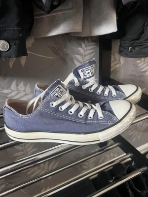 Converse All Star låga sneakers - Klassiska Converse All Star sneakers i lågt snitt. Skorna är mörkblå med vita detaljer, vita snören och tåhätta i gummi. Ovandelen är i canvas och sulan är vit med svart rand. Perfekta för en avslappnad och stilren look.