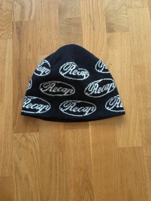 Svart mössa med Recap-logga - Snygg svart mössa från Recap med vit logotyp i ett upprepat mönster runt hela mössan. Klassisk form och mjukt material som sitter skönt på huvudet. Perfekt accessoar för att lyfta din streetwear-look.