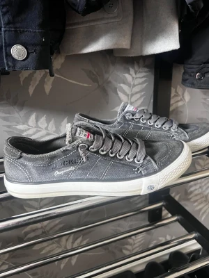 Grå låga sneakers från Dockers - Snygga grå sneakers från Dockers med slitna detaljer och vita sulor. Skorna har klassisk snörning, diskreta sömmar och logotyp på sidan. Perfekta för en avslappnad streetstil och passar till det mesta i garderoben.