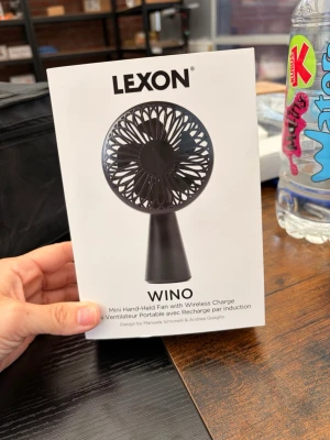 Lexon Wino bärbar fläkt - Säljer en svart Lexon Wino mini handhållen fläkt med trådlös laddning. Fläkten har en rund form med tätt galler och smidig handtag, perfekt att ta med sig när det är varmt. Modern design av Manuela Simonelli & Andrea Quaglio.