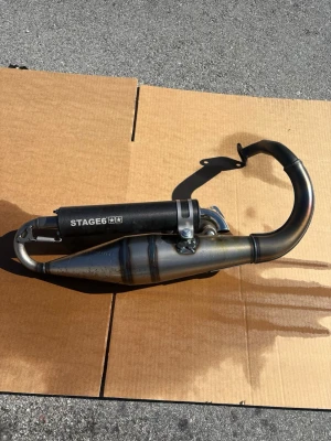Stage6 svart avgassystem scooter - Säljer ett svart avgassystem från Stage6 till scooter eller moped. Utrustad med ljuddämpare och fjädrar, tillverkad i metall med mattsvart finish och synliga svetsfogar. Passar dig som vill uppgradera din moppe med sportigare ljud och look.