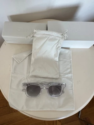 Chimi 04 solglasögon  - Snygga transparenta solglasögon från Chimi/modell 04 med gråtonade linser och fyrkantig form. Glasögonen kommer med vit hård box, vitt skyddsfodral och mjuk påse för förvaring samt en vit putsduk. Perfekt för dig som vill ha en stilren och modern look.