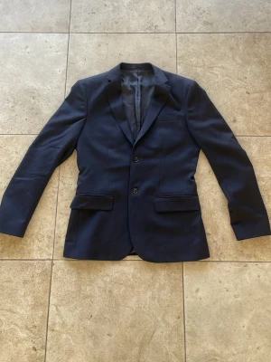 Mörkblå klassisk kavaj - Stilren mörkblå kavaj med två knappar framtill och två fickor med lock. Kavajen har smal passform, klassiska slag och är helfodrad. Perfekt för en dressad look och passar till skjorta eller t-shirt. Materialet känns slätt och lätt.