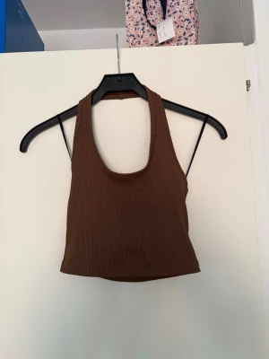 Brun ribbad croppad linnetopp - Snygg brun croppad linnetopp med ribbad struktur och breda axelband. Toppen har en djup urringning fram och öppen rygg, perfekt för varma dagar eller lager på lager. Enkel och stilren design som passar till det mesta.