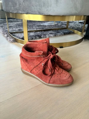 Rostbruna kilklackade sneakers i mocka - Säljer ett par snygga rostbruna sneakers i mocka med kilklack och dekorativa broguemönster. Skorna har snörning framtill, gummisula och en unik design som sticker ut. Perfekta för dig som vill ha något annorlunda och trendigt till din outfit.