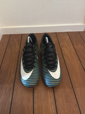 Nike Mercurial fotbollsskor svart/grön - Säljer ett par Nike Mercurial fotbollsskor med svart bas och tunna gröna och blå ränder. Skorna har en stor vit Nike-logga på sidan och ovansidan samt orange dobbar undertill. Snygg, strömlinjeformad design med snörning och syntetmaterial för bra grepp på planen.