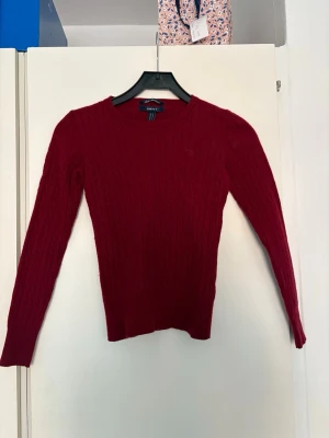 Röd stickad tröja från GANT - Snygg röd stickad tröja från GANT i äkta lammull. Tröjan har rund halsringning, ribbstickade muddar och ett klassiskt kabelstickat mönster. Perfekt för dig som gillar en stilren och tidlös look. Ursprungligen storlek M men skulle säga att den krympt till en större S.
