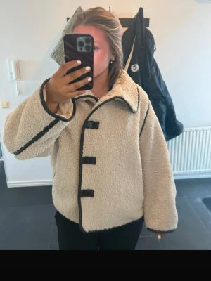 Beige teddyjacka med svarta detaljer - Supermysig beige teddyjacka med svarta kantdetaljer och knäppning framtill. Jackan har hög krage, vida ärmar och är tillverkad i fluffigt teddy-material som håller dig varm. Perfekt för att lyfta din höst- eller vinterstil.